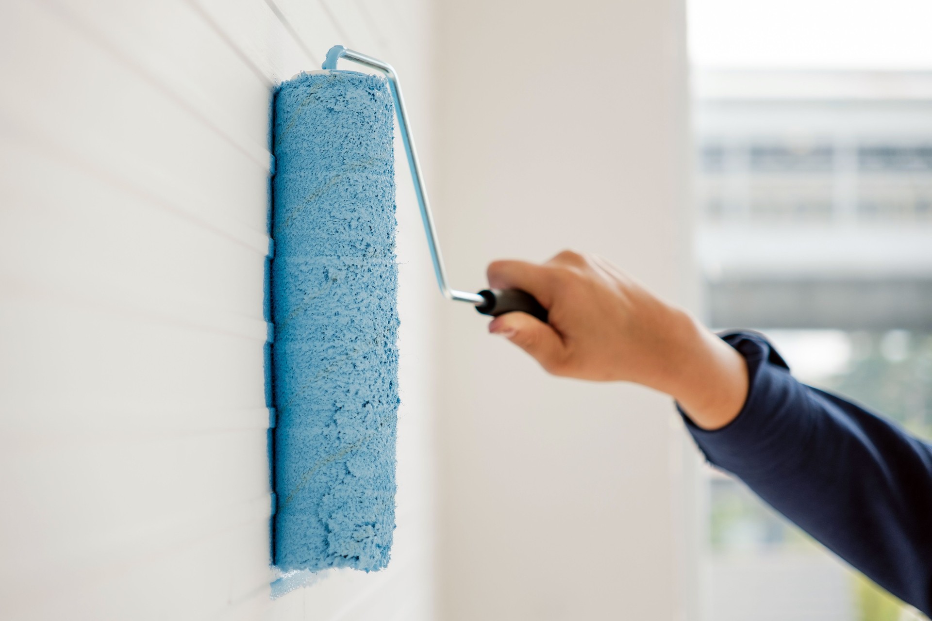 Hand holding a paint roller applying bright blue paint on a white textured wall. DIY home renovation concept, giving a fresh and modern look to interior spaces with bold color.